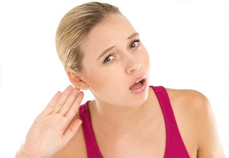 A young blonde woman in a pink tank top holds her hand up to her right ear with a strained, confused expression.